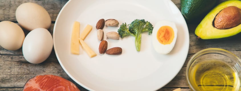 tavola con vari ingredienti che circondano un piatto in cui c'è scritto keto con formaggio, frutta secca broccolo e uovo